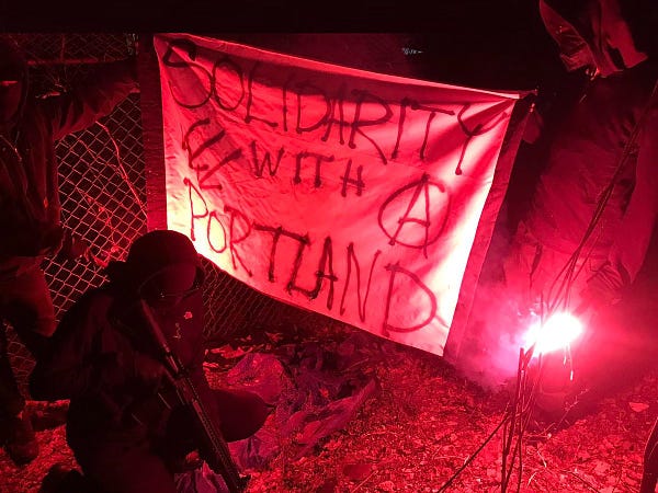 A banner that says “solidarity with portland” and an iron front on one side and an anarchy A on the other.   Some armed comrades are pictured near the banner in bloc