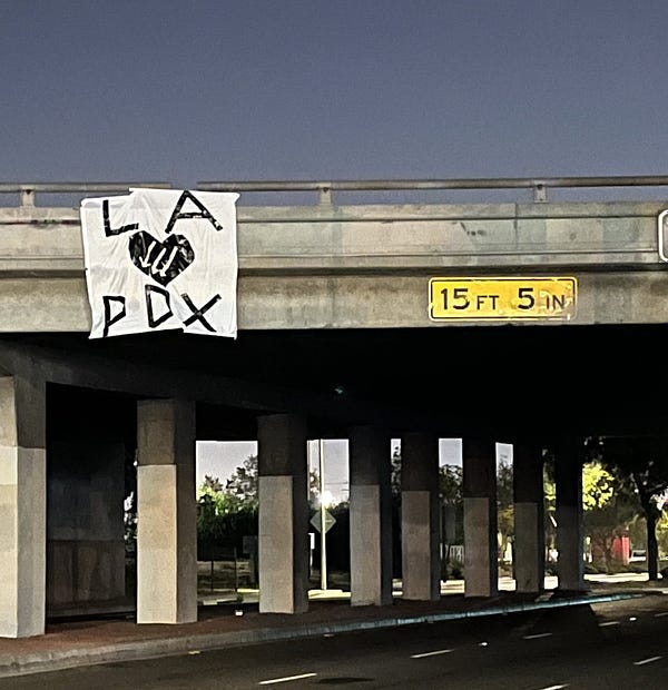 A banner on an overpass that says “LA hearts PDX” in black. The heart is the shape of a heart and filled in with black, and in the center of the heart there is a white iron front.