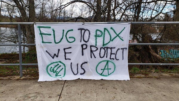 Picture of a sheet banner hanging outside near a river that says "EUG TO PDX - WE PROTECT US" with an antifascists symbol and an anarchy A symbol. 
