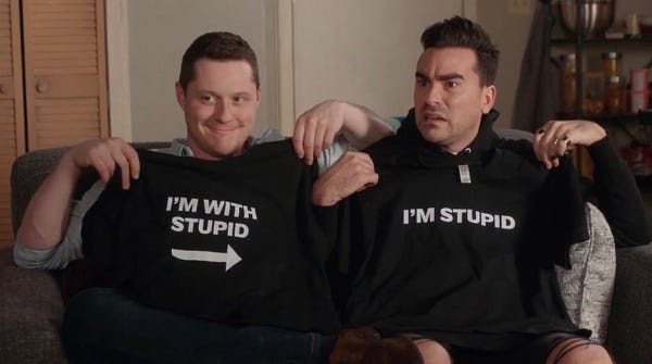 Schitt’s Creek The Bachelor Party. David and Patrick at the apartment. Patrick Brewer looking smug holding up a shirt that says “I’m With Stupid” with and arrow pointing at David. David Rose looking annoyed holding up a shirt that says “I’m Stupid”