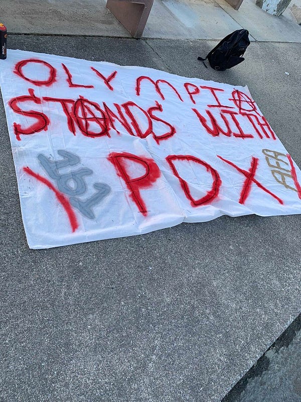A white banner that says “Olympia stands with Pdx” in red writing. The A’s are all anarchy A’s and to the left of the banner it says “161”