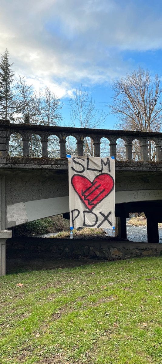 A banner hanging on a bridge that’s all white. The top of it says “SLM” and the bottom “PDX” in black. In the center is a large red heart outlined with black with an iron front in the center