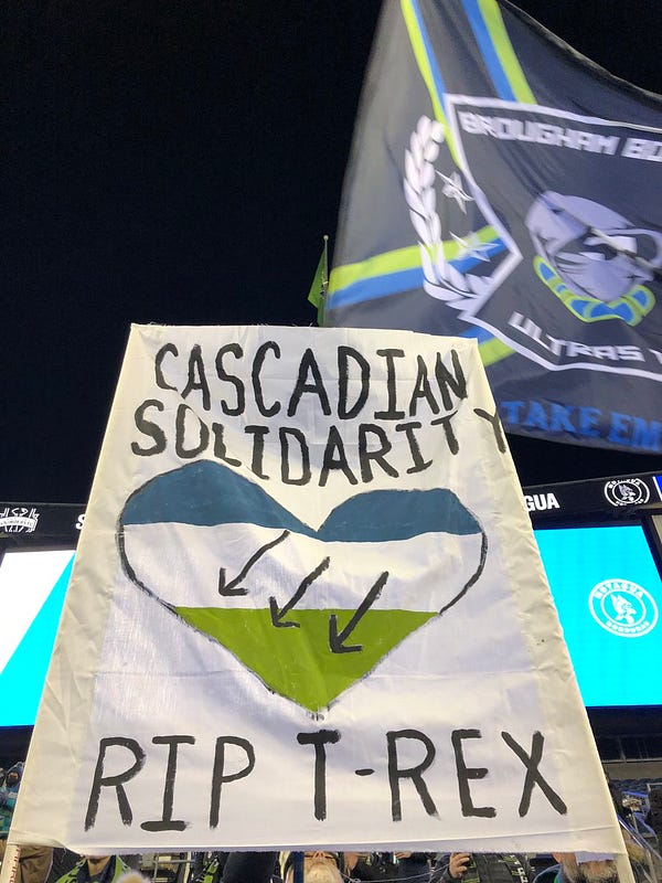 A banner in a soccer stadium reads "Cascadian Solidarity, RIP T-Rex" with a tri-color heart in the Cascadia colors and an Iron Front symbol.