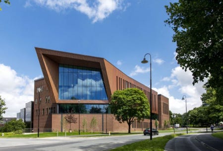 A photograph of the library at the University of Limerick. It is a large, imposing building made from brick and wood. At the nearest end is a huge glass window which spans several floors. The end wall is in the shape of a parallelogram. In the foreground is a tree-lined road and overhead is a bright blue sky dotted with fluffy white clouds 