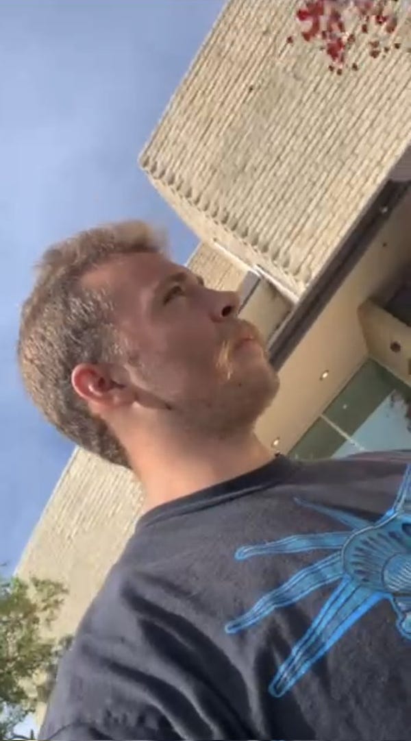 Video still showing the profile of a white man with a short, light brown haircut and light brown mustache wearing a black T-shirt with some kind of design in blue printed on the front. Behind him is a tan brick building. Above him is blue sky.