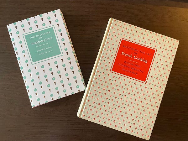 Two books on a table. The left-hand book is an aqua and brown print on a white background. The right-hand books is a bit larger, and is a red and blue print on a faded, off-white background. The prints and titles on the two books are intentionally similar. The left-hand book is “Chocolate Cake for Imaginary Lives” by Genevieve Jenner. The right-hand book is “Mastering the Art of French Cooking” by Julia Child, Louisette Bertholle, and Simone Beck.