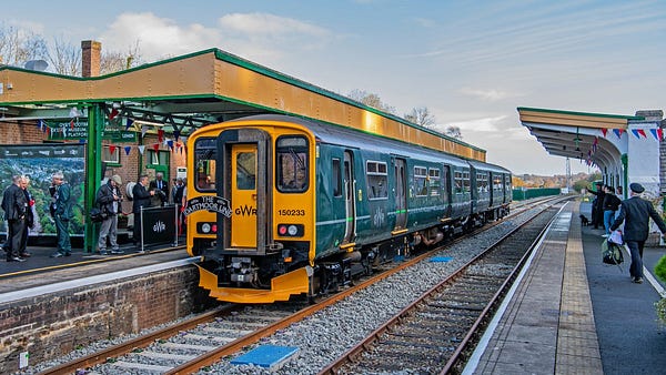 A train waits at a station on the Dartmoor Line, with passengers on the platform and bunting hanging from the waiting areas.