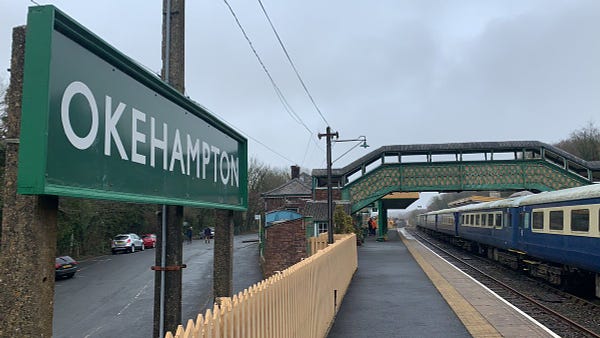 Okehampton station sign on the platform with a footbridge and train in the background.