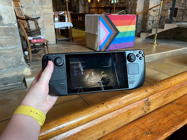 A Steam Deck, playing the Game Elden Ring, with the character sat at a save point (known as a “Site of Grace”, held above a church pew in front of an altar with the progress pride flag draped over it.