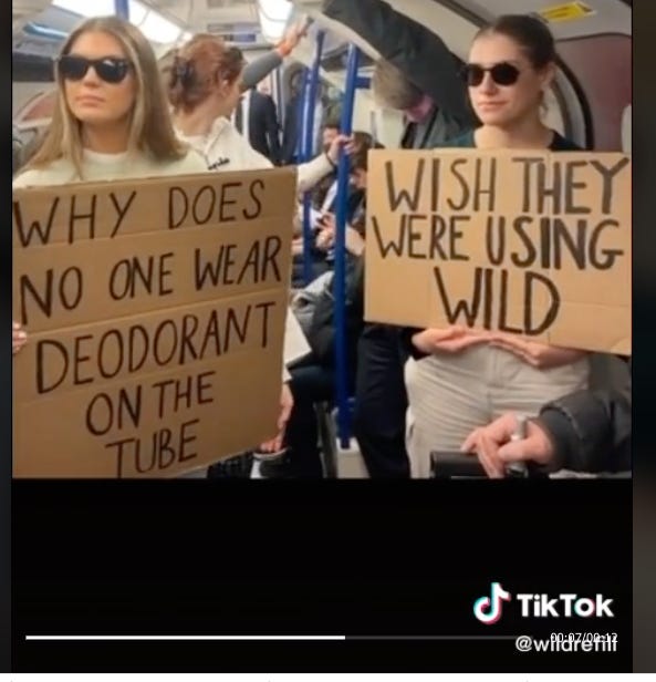 two people holding cardboard signs on the tube saying “why does no one wear deoderant on the tube” and “wish they were using wild"