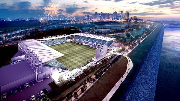 Another aerial view image render of the entire stadium taken from the perspective of a drone over the river. It is facing the open side of the stadium and shows fireworks exploding overhead. Downtown Kansas City lies in the near distance just to the right of the stadium.