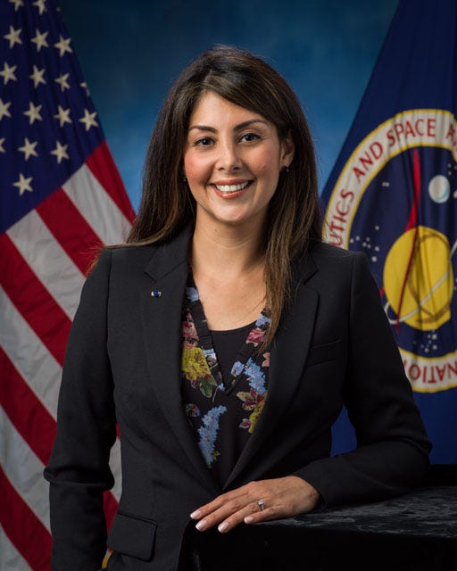 Retrato oficial de Diana Trujillo, una mujer de cabello oscuro, largo y lacio, con ojos oscuros y traje chaqueta, antes las banderas de Estados Unidos y la NASA.