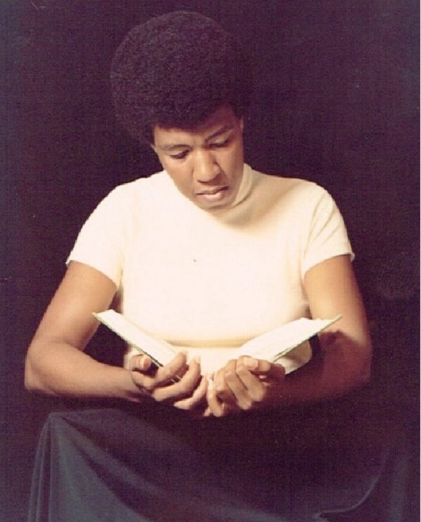 Octavia Butler sits against a black background wearing a white top and what looks like a black skirt. Her head is bowed, reading a book that is perched on her lap.