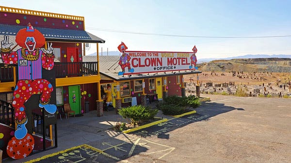 photo of the outside of a small motel. big clown signs are outside of it and a sign above reads “welcome to the world renown clown motel”. you can see there is a cemetery less than like 10 feet away from it.