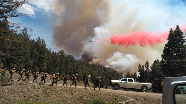 A fire hand crew heads walks on a dirt road while a plane in the background drops retardant over a smoke column.