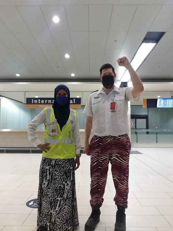 A female and male airport security screening officer standing inside an airport.