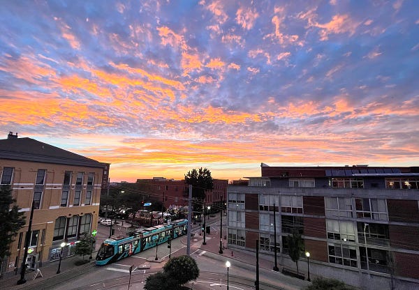 View of Delaware Street in Kansas City’s River Market neighborhood, with a brilliant yellow, pink and blue sunset overhead and a KC Current wrapped Streetcar passing by
