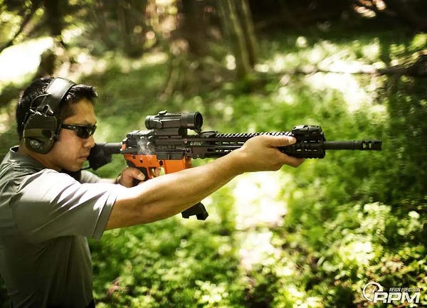 Officer Christopher Brownlee pointing a tactical rifle in the woods in a vest, sunglasses, and ear protection.