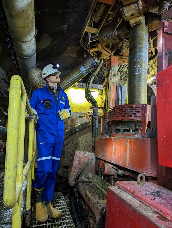 A lucky guy in a blue jumpsuit and hardhat stares at a big machine in a mine. The machine has a pipe about 30 centimeters in diameter ascending into the top of the tunnel above.