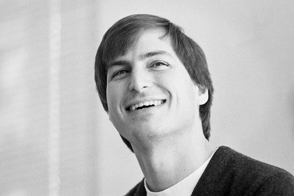 A black and white photo of Steve Jobs glancing upward with a wide smile.