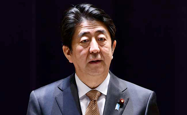 Shinzo Abe wears a suit and stands under a light in front of a dark background