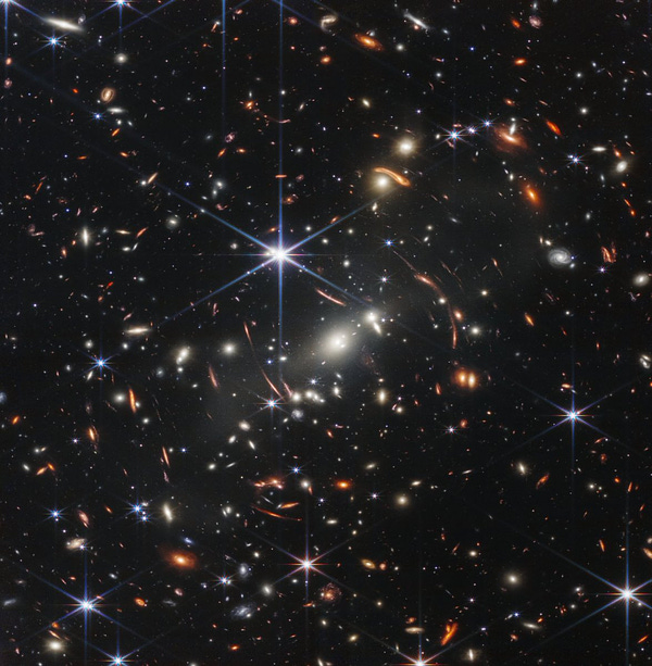 The background of space is black. Thousands of galaxies appear all across the view. Their shapes and colors vary. Some are various shades of orange, others are white. Most stars appear blue, and are sometimes as large as more distant galaxies that appear next to them. A very bright star is just above and left of center. It has eight bright blue, long diffraction spikes. Between 4 o'clock and 6 o'clock in its spikes are several very bright galaxies. A group of three are in the middle, and two are closer to 4 o'clock. These galaxies are part of the galaxy cluster SMACS 0723, and they are warping the appearances of galaxies seen around them. Long orange arcs appear at left and right toward the center.