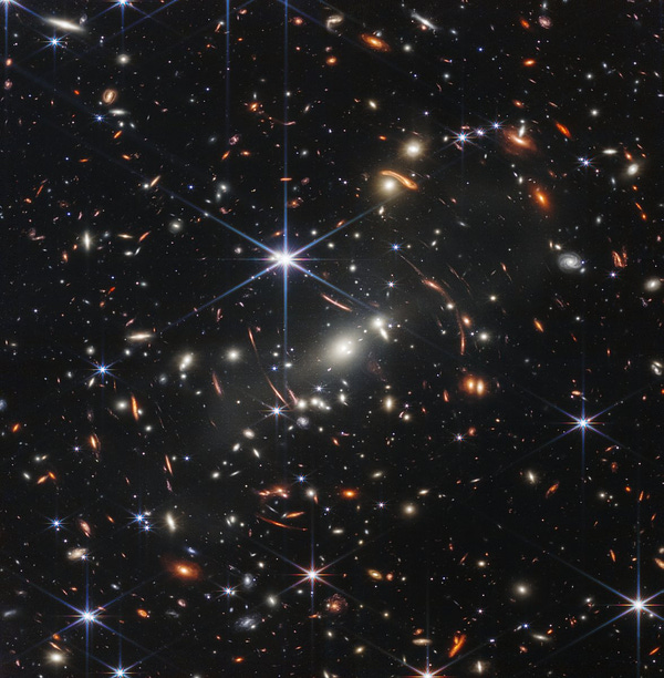 The background of space is black. Thousands of galaxies appear all across the view. Their shapes and colors vary. Some are various shades of orange, others are white. Most stars appear blue, and are sometimes as large as more distant galaxies that appear next to them. A very bright star is just above and left of center. It has eight bright blue, long diffraction spikes. Between 4 o’clock and 6 o’clock in its spikes are several very bright galaxies. A group of three are in the middle, and two are closer to 4 o’clock. These galaxies are part of the galaxy cluster SMACS 0723, and they are warping the appearances of galaxies seen around them. Long orange arcs appear at left and right toward the center.
