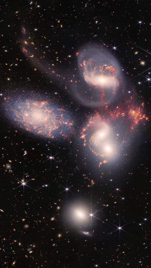 A group of five galaxies that appear close to each other in the sky: two in the middle, one toward the top, one to the upper left, and one toward the bottom. Four of the five appear to be touching. One is somewhat separated. In the image, the galaxies are large relative to the hundreds of much smaller (more distant) galaxies in the background. All five galaxies have bright white cores. Each has a slightly different size, shape, structure, and coloring. Scattered across the image, in front of the galaxies are number of foreground stars with diffraction spikes: bright white points, each with eight bright lines radiating out from the center. 