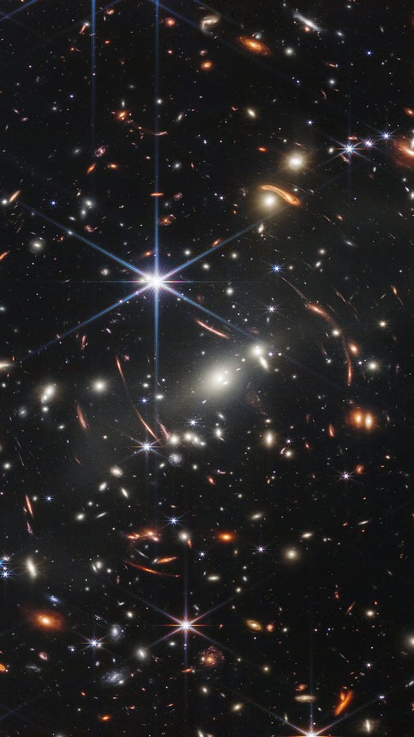 The background of space is black. Thousands of galaxies appear all across the view. Their shapes and colors vary. Some are various shades of orange, others are white. Most stars appear blue, and are sometimes as large as more distant galaxies that appear next to them. A very bright star is just above and left of center. It has eight bright blue, long diffraction spikes. Between 4 o’clock and 6 o’clock in its spikes are several very bright galaxies. A group of three are in the middle, and two are closer to 4 o’clock. These galaxies are part of the galaxy cluster SMACS 0723, and they are warping the appearances of galaxies seen around them. Long orange arcs appear at left and right toward the center.