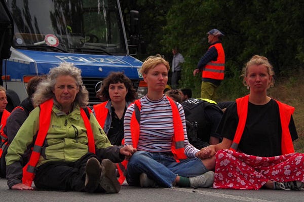 Janine O‘Keefe, Lea Bonasera sowie Lisa und Maja Winkelmann in orangenen Warnwesten auf der Straße sitzend während einer Autobahn-Blockade. Im Hintergrund ein Polizeifahrzeug.