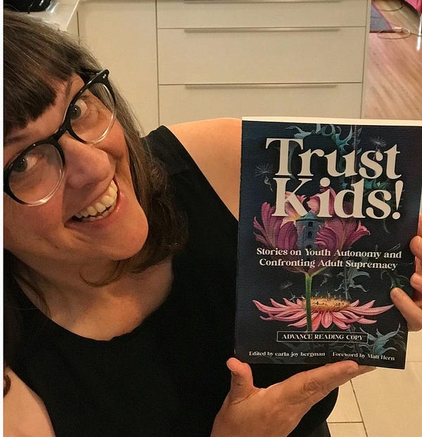 a colour photo of carla joy bergman smiling as they hold an advanced reading copy of Trust Kids! -- her new book! 