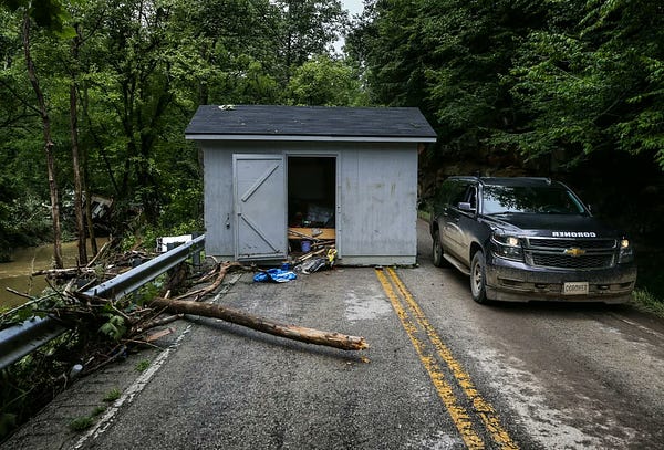 A small grey shed is washed atop a narrow highway as a black Chevrolet SUV belonging to the Knott County Coroner drives past it.  The road is on a mountain bend from Troublesome Creek outside Fisty, Kentucky. (Photo Credit: Matt Stone/Courier Journal)