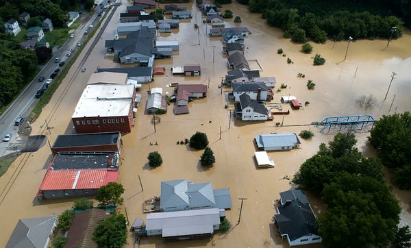 This image is a flyover shot of the town of Garrett in Floyd County, Kentucky.  Brown floodwater can be seen covering the town and buildings are partially submerged in the brown floodwater.  We can see several commercial and residential buildings as well as a blue bridge to the right and a small two-lane road to the left. (Photo credit: Matt Stone/Courier Journal)