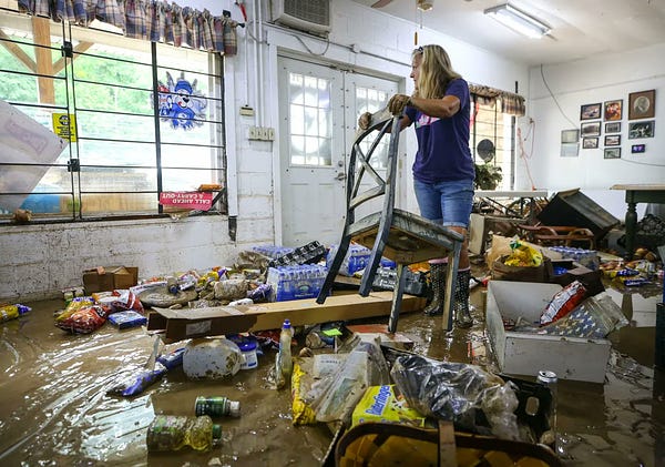 A woman can be seen lifting a flood-damaged chair inside of a building. 
 The building is completely flood damaged with the floor caked in mud in 
 flood water.  The build is Campbell's Grocery store in Garrett Kentucky. 
 Various grocery store items are strewn about on the floor, caked in mud as light shines into the building through a large glass window off to the left. From the article: "I've never seen it this bad," said Campbell's Grocery employee Kella Sloan, as she helps clean up the muddy mess that Thursday's flood left in Garrett, (Photo Credit: Matt Stone/Courier Journal)