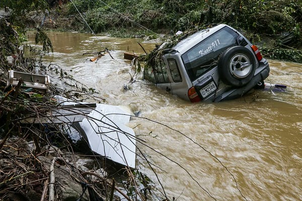 A small grey SUV bearing a "Knott 911" sign on the back liftgate glass is seen partially submerged in a creek.  The creek has brown muddy water flowing swifty through it as white caps can be seen around the vehicle, with debris on the sides of the creek.  The creek pictured is the Right Fork Troublesome Creek in Hindman, Kentucky. (Photo credit: Matt Stone/Courier Journal)
