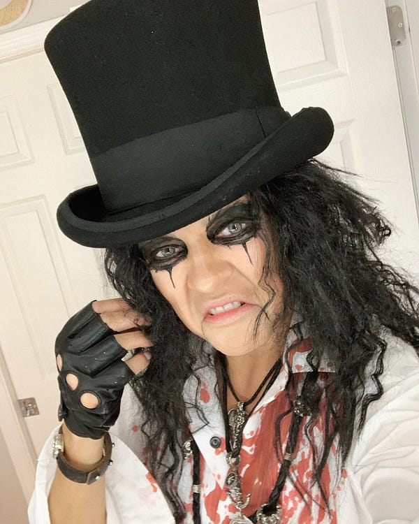 Photo showing Kari Lake wearing a tall black top hat, a black wig, thick black eye shadow, a white shirt covered in fake blood, a leather biker glove, and various necklaces. She appears to indoors with a white door behind her. 