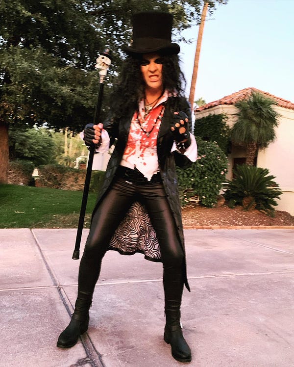 Photo showing Kari Lake wearing a tall black top hat, a black wig, thick black eye shadow, a white shirt covered in fake blood, a long black jacket, tight black leather pants, a leather biker glove, and various necklaces. She is holding a black cane topped with a small white skull. She is standing outdoors on a slab of concrete. Trees, grass, bushes and a house can be seen behind her. 