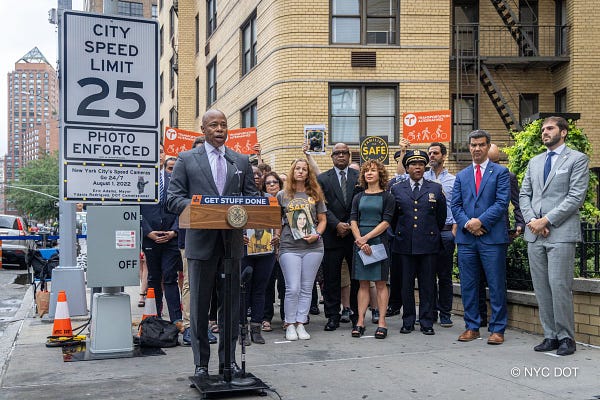 Mayor Adams speaks at a lectern. There is a group standing behind him, including Commissioner Rodriguez. Behind the mayor is a sign that says city speed limit 25. Photo Enforced. New York city's speed cameras go 24/7 August 1, 2022. Eric Adams, Mayor. Ydanis Rodriguez, D O T commissioner. There is an on off switch below the sign.