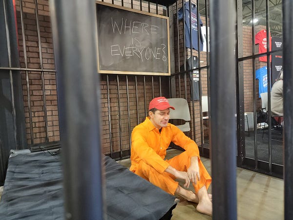 A man in an orange jumpsuit and MAGA hat sits on the floor in a jail cell visibly weeping. Next to him, a jailhouse cot. Above his head, a blackboard that says "where is everyone?" in chalk