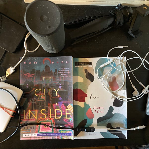 A very messy black desk full of cables, external hard drives, headphones, Bluetooth speakers, iPhone tripods... and two paperback books side-by-side at the bottom: Samit Basu's THE CITY INSIDE and Joma West's FACE.