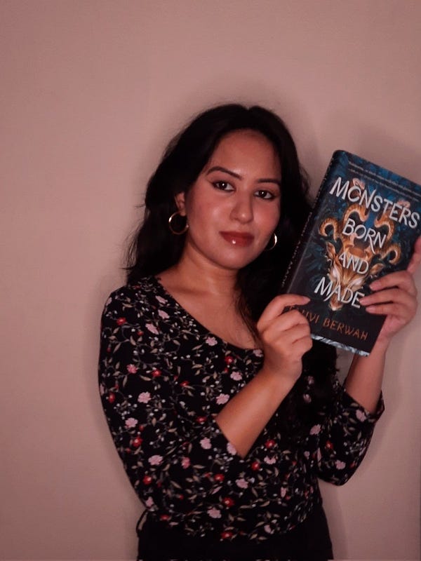 debut author tanvi berwah holds up a copy of her fantasy book, Monsters Born and Made. Tanvi is standing before a pink wall. She's wearing a black top with a flower print and gold earrings.