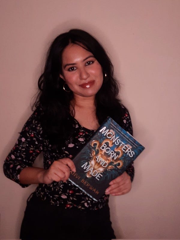 debut author tanvi berwah holds up a copy of her fantasy book, Monsters Born and Made. Tanvi is standing before a pink wall. She's wearing a black top with a flower print and gold earrings.