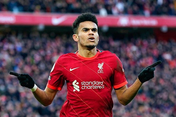 Luis Díaz corre com os braços abertos comemorando um gol do Liverpool. Ele veste luvas e a camisa vermelha do clube, com o nome "Standard Chartered" nela