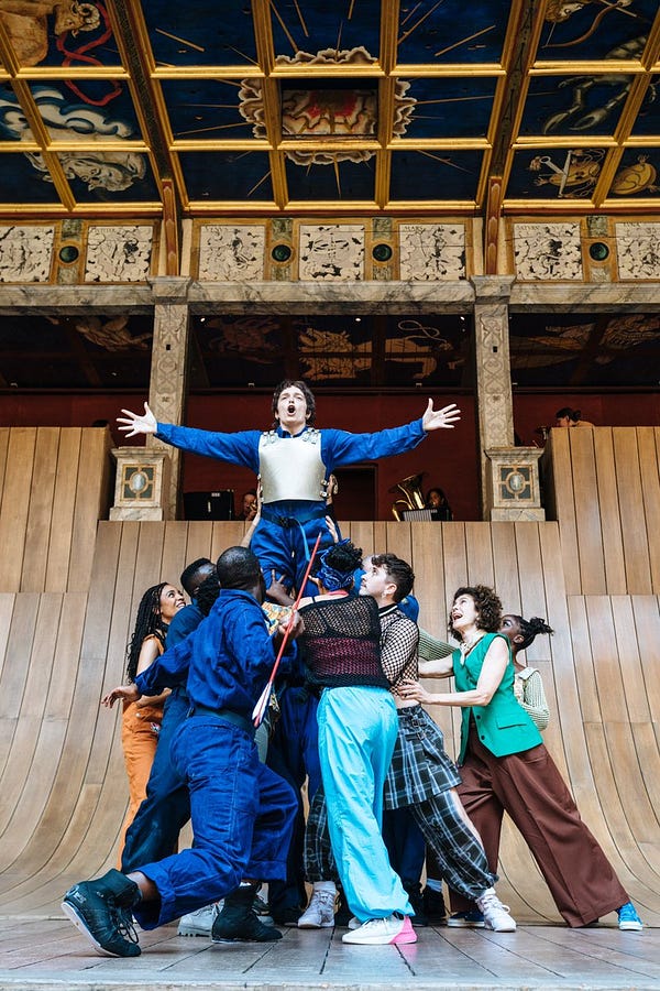 An actor wearing a blue boiler suit and silver chest plate is held aloft in the air on the shoulders of a group of other actors. Their arms are spread wide and they are shouting passionately. The Globe stage is covered with a curved wooden ramp.