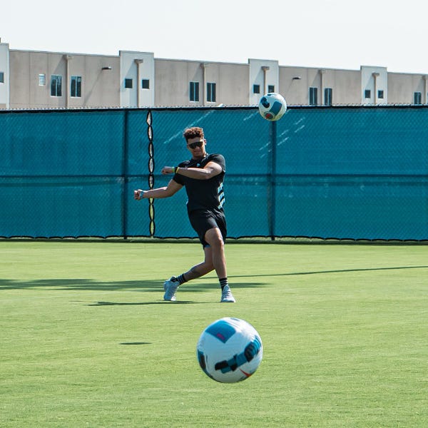 Patrick Mahomes kicking a soccer ball with ball mid-air. He is wearing all black with sunglasses and leaning back in stance.