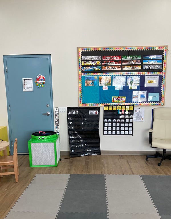 Classroom with white walls, blue door, brown wood floor and foam block flooring. A bulletin board hangs on the wall with months and colours. Green coloured air filter with googly eyes sits on floor