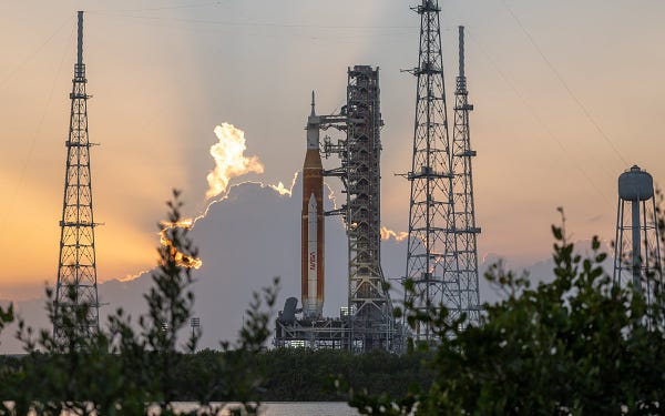 The Space Launch System rocket sits on the launch pad. In the background, the sun rises behind clouds. Green bushes sit in the foreground. 