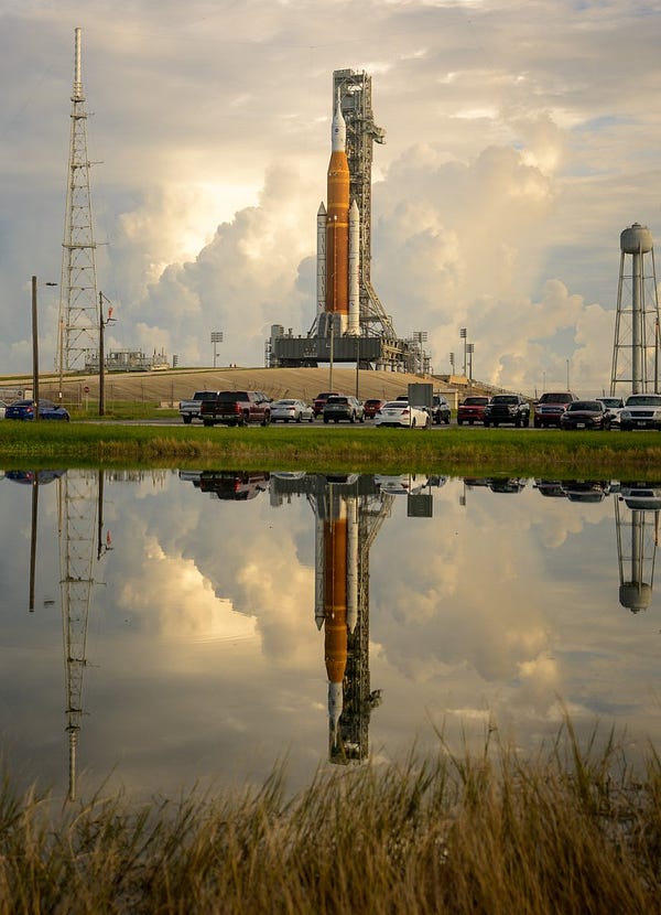 The Space Launch System rocket stands upright on the launchpad. The background is the late afternoon sky dominated by towering clouds. The rocket has an orange central fuel tank with two white rocket boosters on either side. The rocket is reflected in the water in the foreground of the image, framed by marsh grass. Between the water and the rocket is a parking lot with over a dozen parked cars. Photo credit: NASA/Bill Ingalls