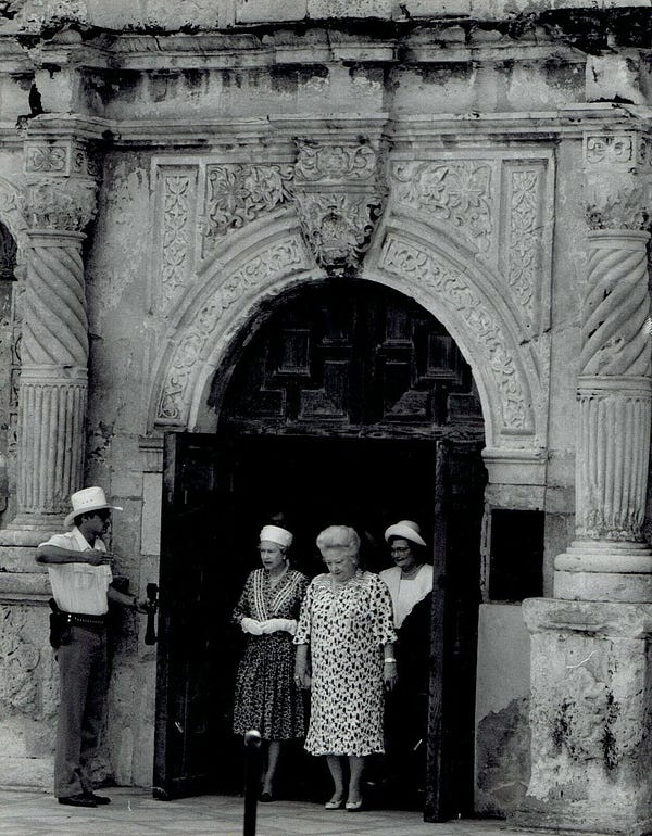 Queen Elizabeth exits the distinctive entryway of the Alamo with a woman beside her and another following; a guard stands at the door.