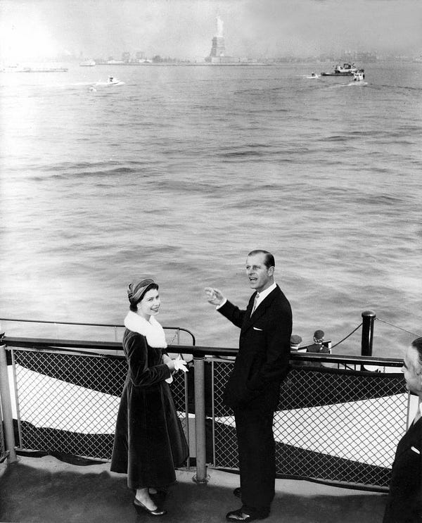 A black and white photograph of Queen Elizabeth II and Prince Philip on a boat overlooking the Hudson River with the Statue of Liberty in the background.